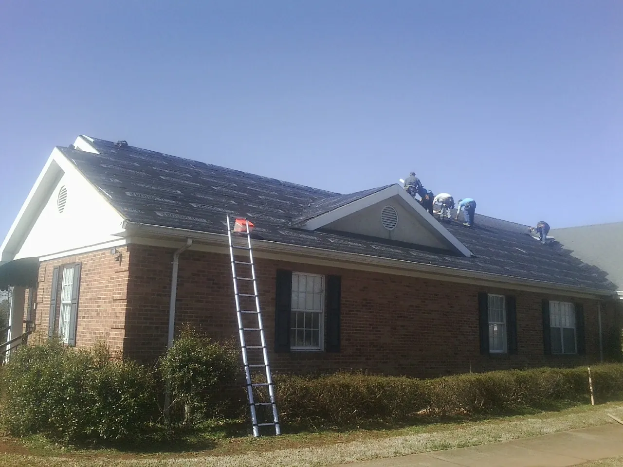 Crew tearing shingles off a brick ranch roof with a ladder set — Metal Roof Repair work by our crew in Farrell