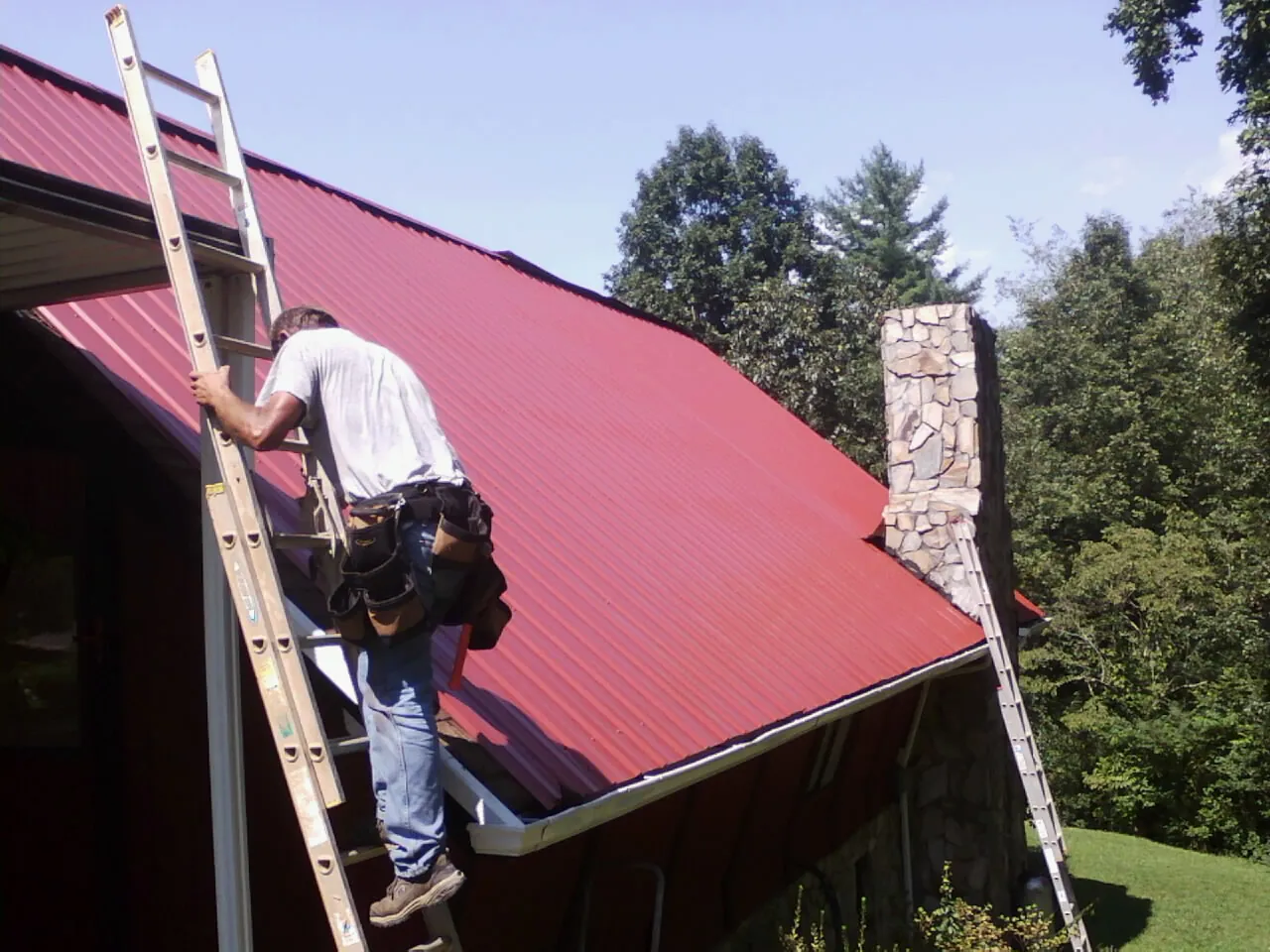 Roofer on a ladder beside a red metal roof and stone chimney — Soffit & Fascia Repair by Roofing Brothers in Farrell
